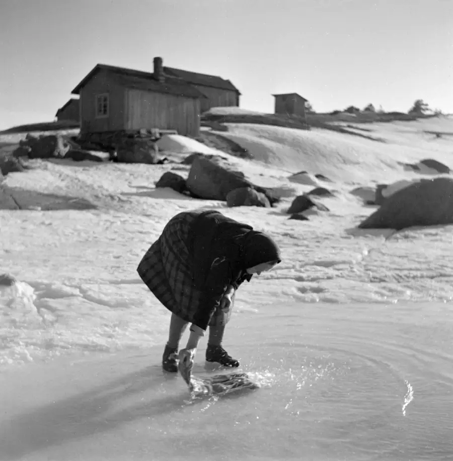 A person in winter clothing bends down on a snowy, icy surface, possibly washing something in a hole in the ice. Wooden houses and boulders are visible on a snowy hill in the background.