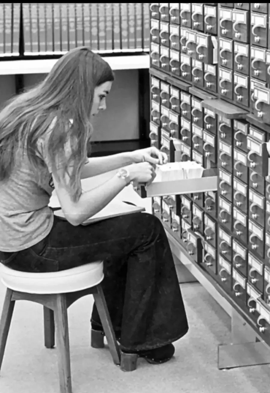 A person sits on a stool and searches through a card catalog drawer in a library, surrounded by rows of small drawers and bookshelves.