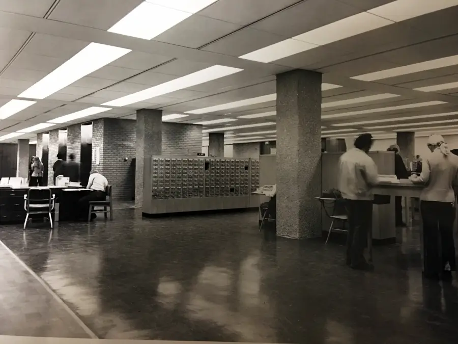 Black-and-white photo of a spacious library interior with card catalog drawers, desks, and several people reading or working. The ceiling has recessed lighting, and the floor is shiny and reflective.