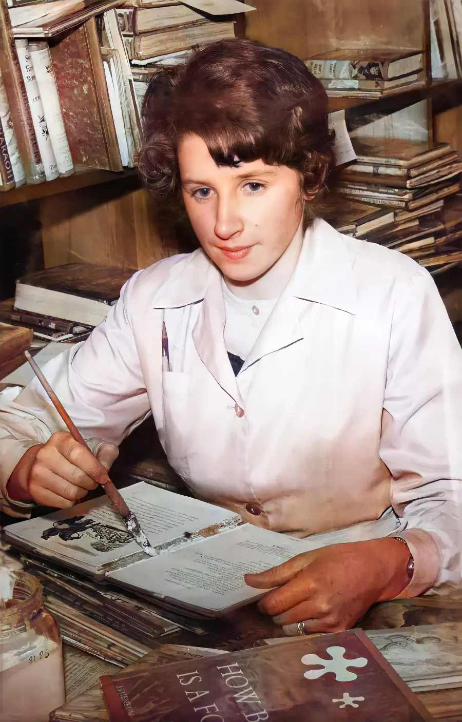 A woman in a white lab coat sits at a desk with books, writing or drawing in an open book. Shelves filled with more books are in the background. She looks focused and is holding a brush or pen.