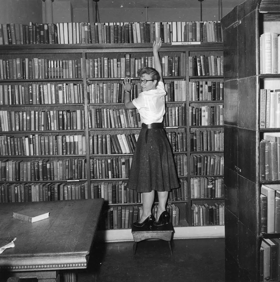 A woman in a skirt and glasses stands on a stool, reaching for a book on a tall, crowded bookshelf in a library. A table with books is in the foreground.