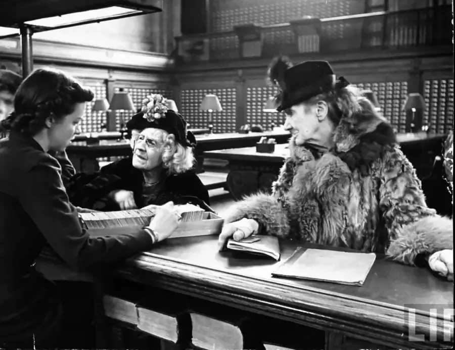 Two elderly women in ornate hats and fur coats speak with a librarian across a counter in a large library, surrounded by books and lamps.