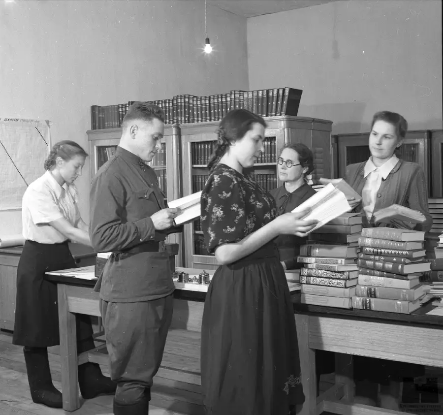 Five people stand and read or sort books in a library or office, with bookshelves and stacks of books on tables. The room is well-lit and the people appear focused on their tasks.