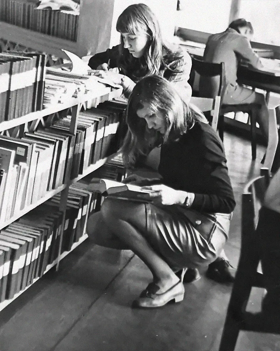 Two young women look at books on a low library shelf, one standing and one kneeling. In the background, a man reads at a table. The scene appears to be in a library or study area.