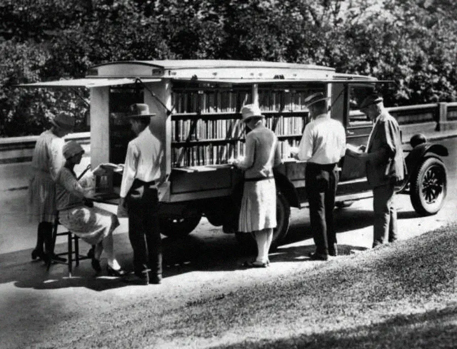 A black-and-white photo of people browsing books at a bookmobile, which is a truck with shelves of books and a table outside. The scene appears to be from the early 20th century.