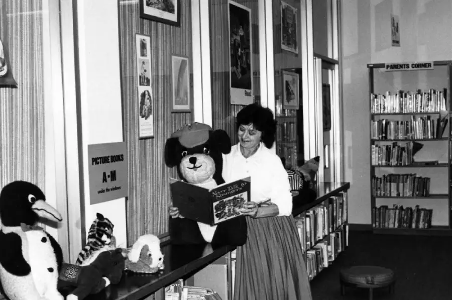 A woman in a library smiles while reading a book to a large stuffed bear wearing a hat. Plush toys line the counter, and bookshelves are visible in the background.