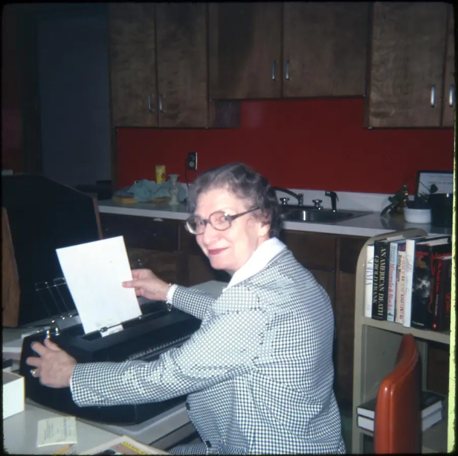 An older woman in glasses and a checked blazer sits at a desk, smiling and placing paper into a typewriter. Books, a chair, and a kitchen area with a red wall and wooden cabinets are visible in the background.