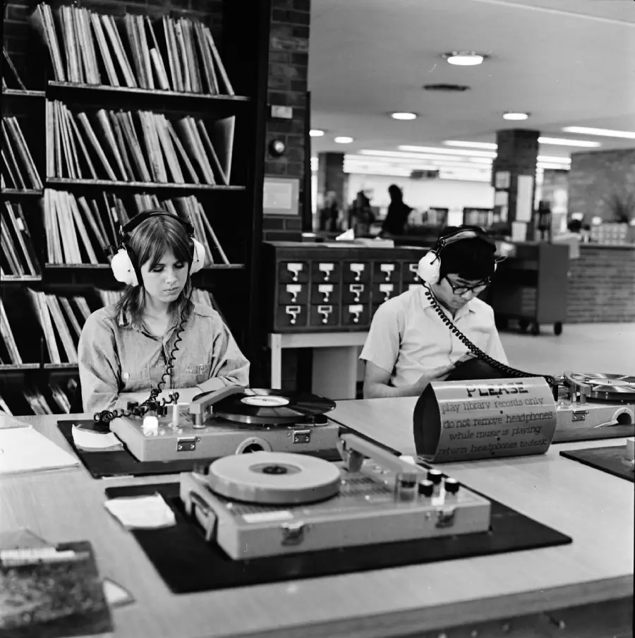 Two people wearing headphones sit at a library table listening to vinyl records on turntables. Shelves of records and a card catalog are visible behind them. The setting appears to be a public or university library.