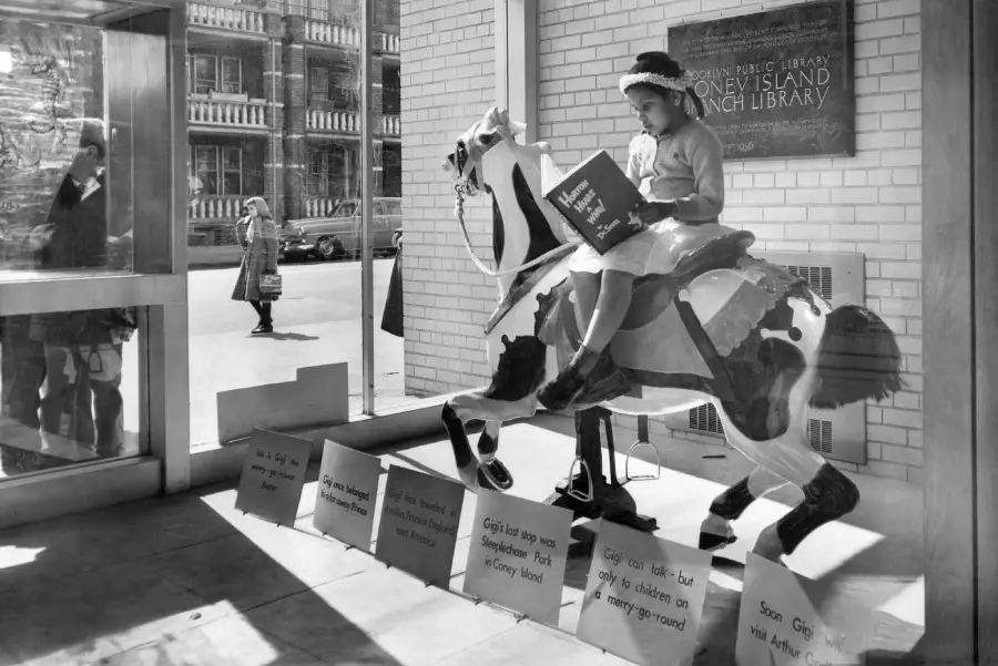 A young girl in a dress and hat sits on a carousel horse indoors, reading a book. Signs with text are displayed below. A library plaque and windows showing people outside are visible in the background.