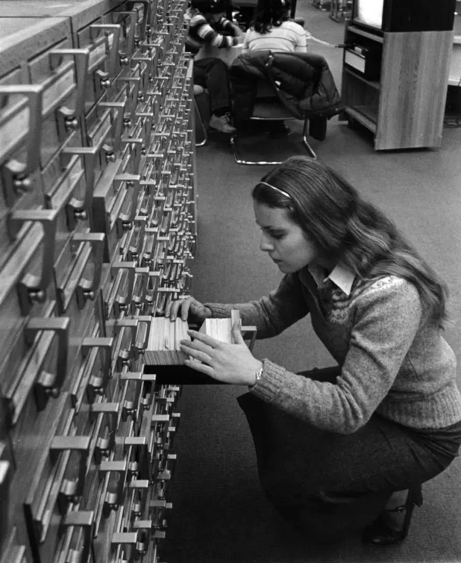 A woman kneels and looks through a card catalog drawer in a library. Shelves of drawers run alongside her, and other people are seated in the background.