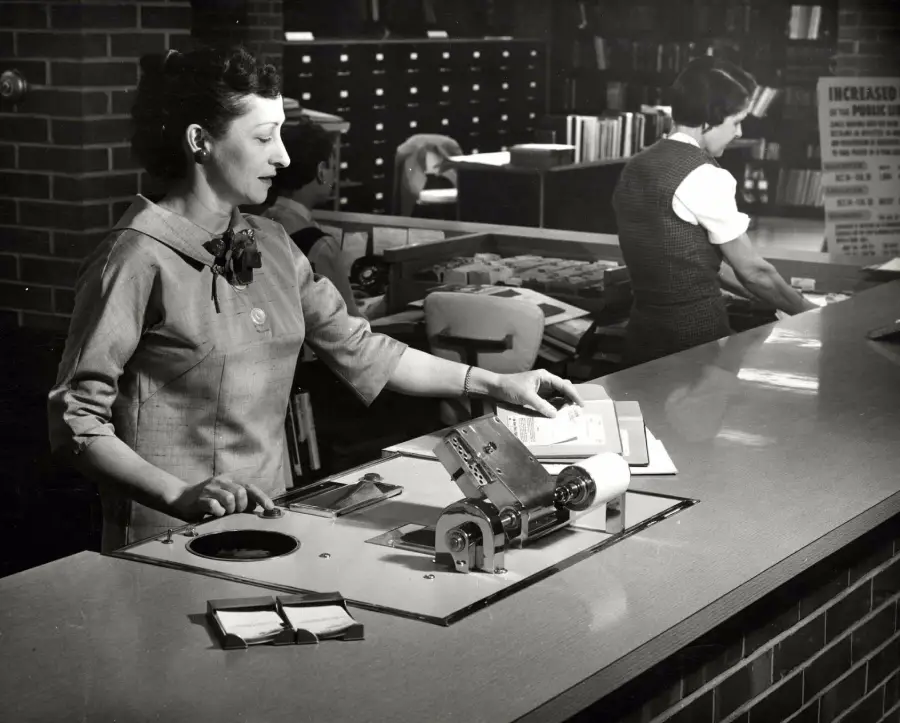 A woman uses a mechanical device at a library counter while another woman works in the background near bookshelves and filing cabinets. The setting appears to be a mid-20th-century library.