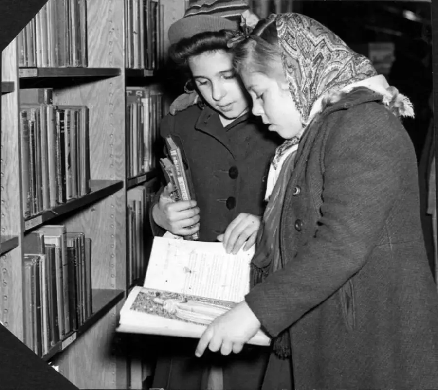 Two girls in coats and scarves stand by bookshelves, one holding books and the other showing her an open book. They appear to be reading or discussing the contents together in a library or bookstore.