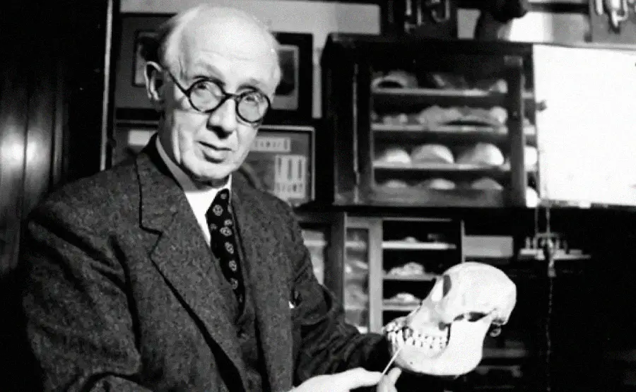 An older man wearing glasses and a suit holds a skull model in a room filled with shelves and specimen cases, suggesting a scientific or anatomical study setting.