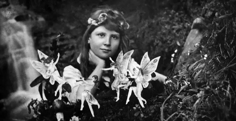 A vintage photo of a girl with a flower crown posing outdoors, surrounded by cut-out paper fairies, blending fantasy and reality in a garden setting.