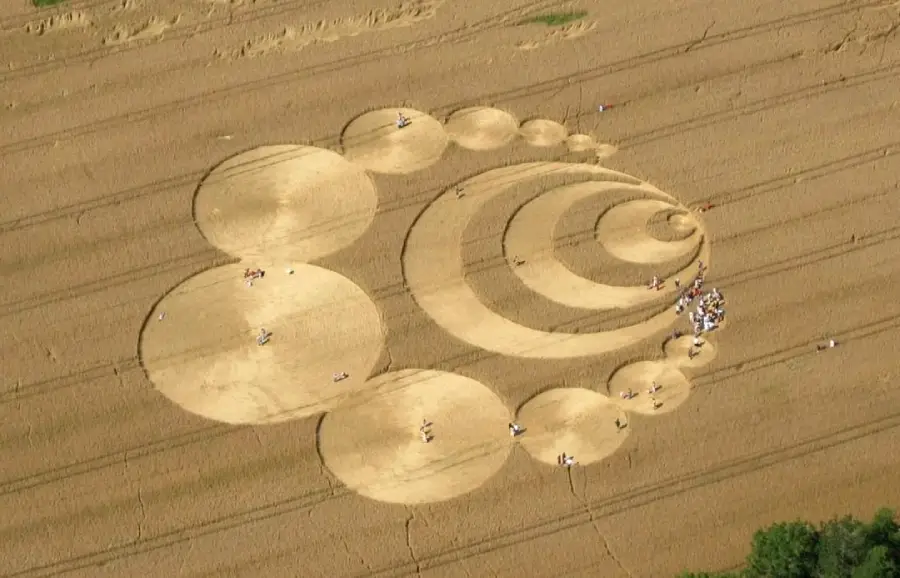Aerial view of a large crop circle pattern featuring multiple interconnected circles and spirals in a golden wheat field, with small groups of people walking around inside and near the design.