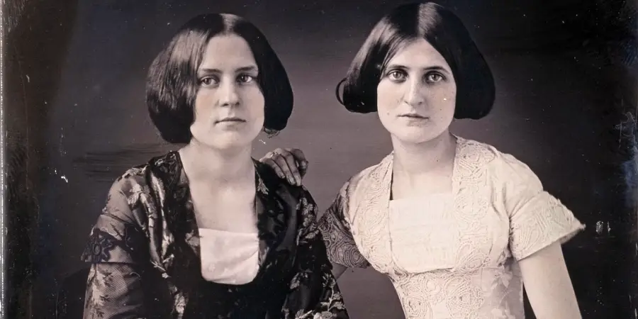 Two women with bobbed dark hair wearing 19th-century dresses sit side by side, one resting her hand on the other's shoulder, both gazing directly at the camera with serious expressions.