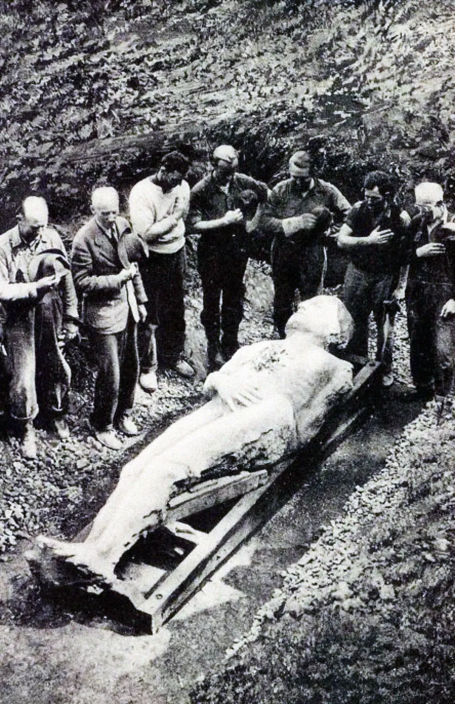 A group of people stand around a large stone statue lying on its back outdoors, with some bowing their heads or holding their hands to their chests in a solemn manner.
