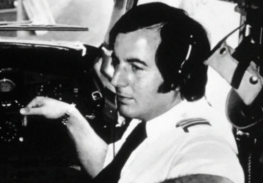 Black and white photo of a male pilot wearing a headset and uniform inside an airplane cockpit, with one hand on the controls and instrument panels visible in the background.