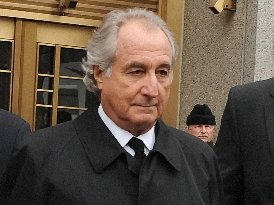 An older man with gray hair, wearing a black coat and tie, stands outside a building with gold-framed doors. There are other people partially visible in the background.