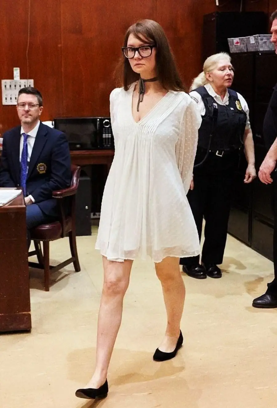 A woman in a white dress and black flats, with hands behind her back, walks in a courtroom. She has long brown hair and glasses. Police and others are present in the background.