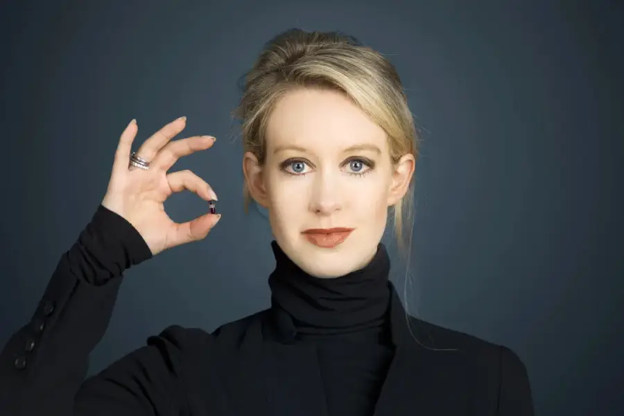 A woman with blonde hair in a black turtleneck holds up a small object between her fingers, making an “OK” gesture, and looks directly at the camera against a plain dark background.