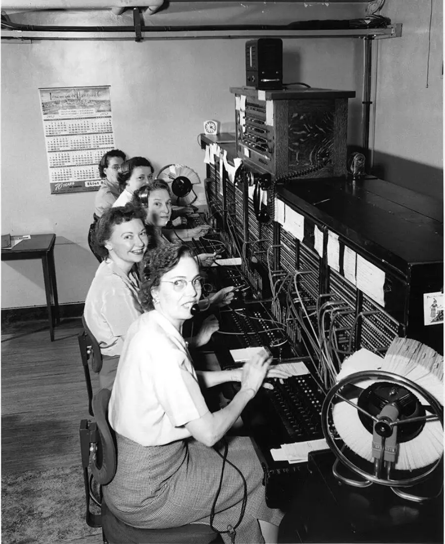 Five women sit in a row at a vintage telephone switchboard, connecting calls with cords and plugs. A wall calendar hangs behind them. The setting appears to be an office from the mid-20th century.