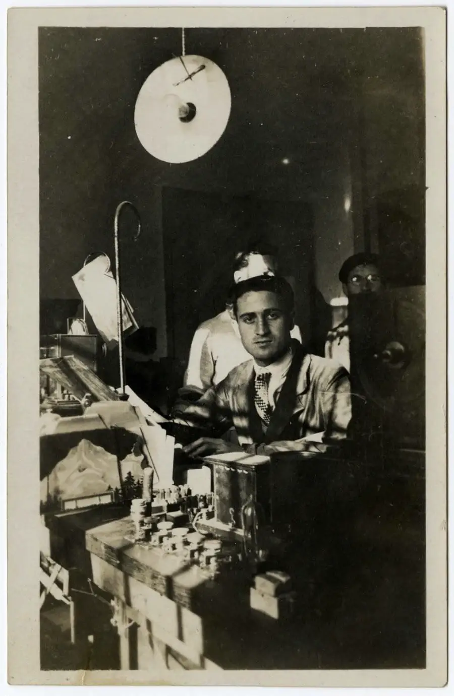 A man in a lab coat sits at a cluttered workspace with scientific equipment. A large lamp hangs above him, and two people stand in the background, partially obscured. The scene appears to be an old laboratory.