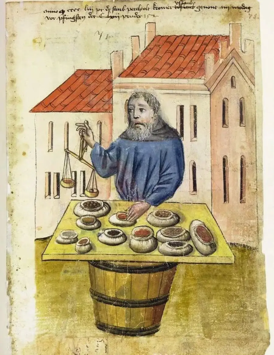A medieval merchant with a gray beard stands behind a table displaying various cheeses, using a balance scale. Buildings with red roofs are visible in the background.