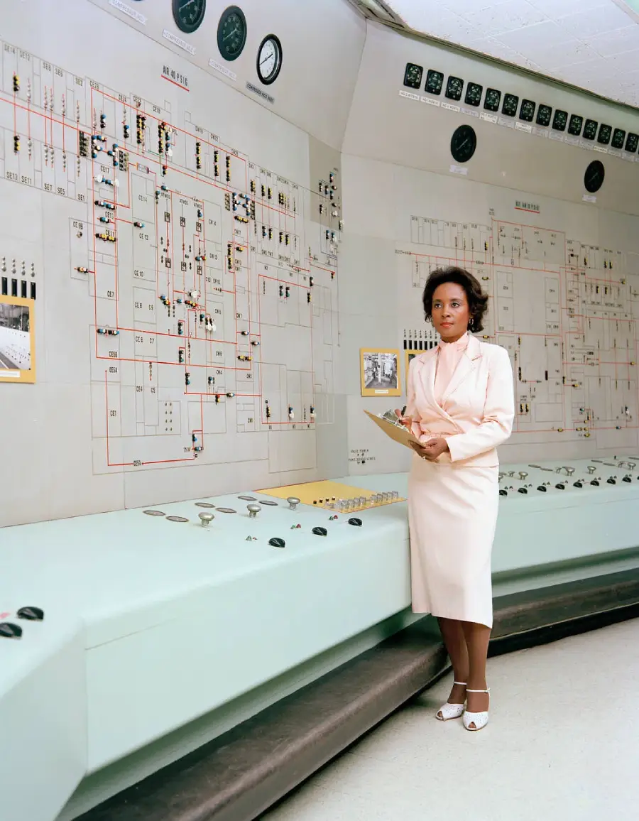 A woman in a light pink dress stands with a clipboard in front of a large control panel covered in buttons, gauges, and diagrams.