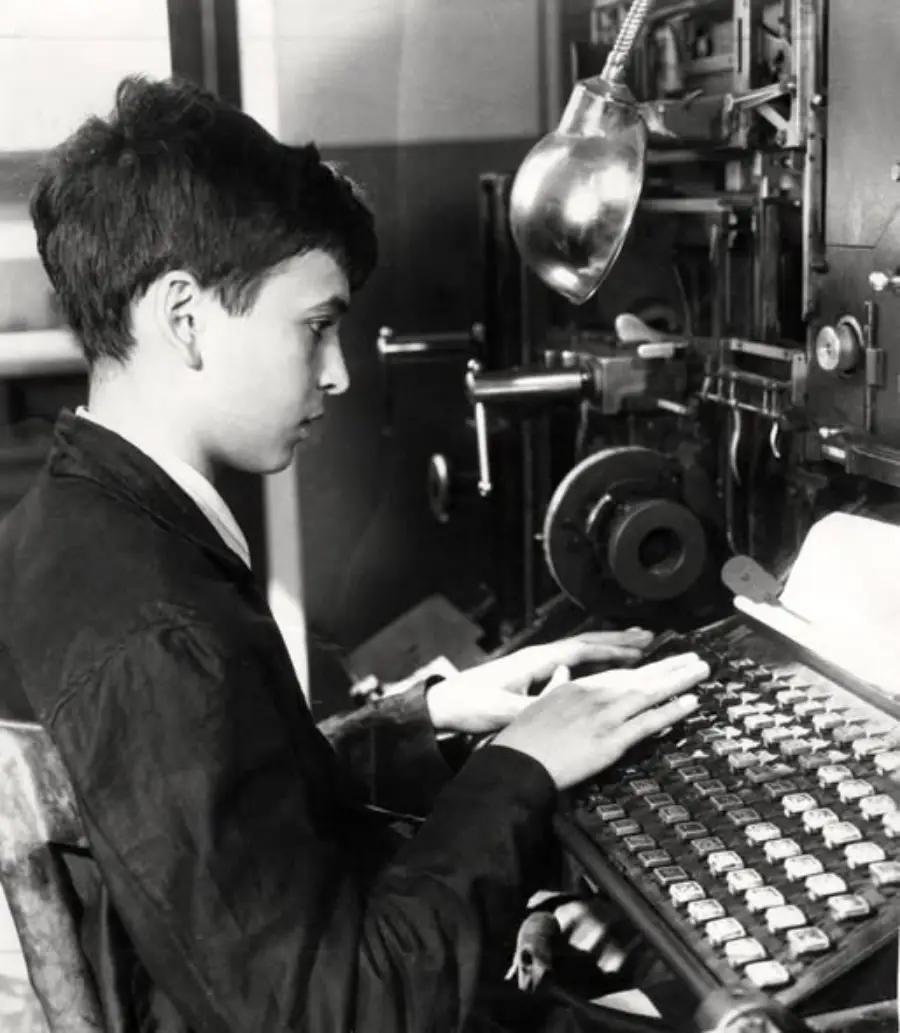 A young person sits at an old-fashioned typesetting machine, looking focused while typing on a large keyboard. A lamp shines overhead, illuminating the workspace filled with mechanical parts.