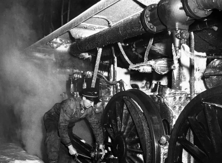 A worker in overalls and a cap repairs the wheel area of a large steam locomotive at night, surrounded by steam and snow, using a tool.
