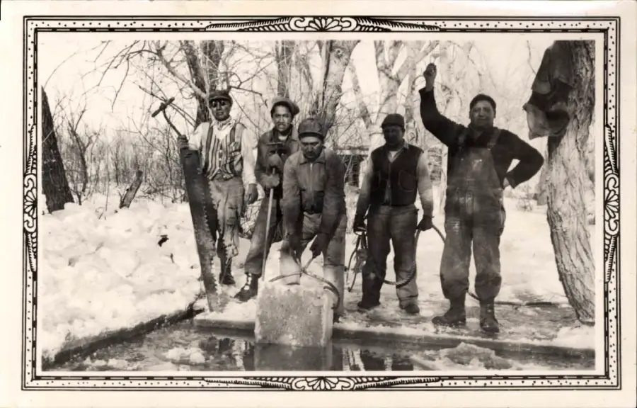 Five men pose outdoors in winter by a hole in the ice. One holds an ice saw, two hold tools, and another lifts a block of ice. Snow-covered ground and bare trees are in the background.