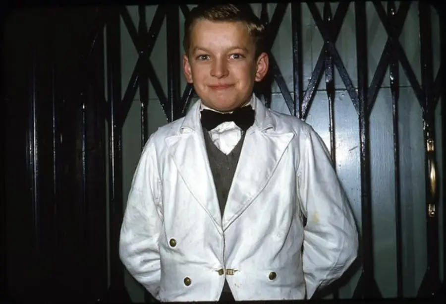 A young boy in a white double-breasted suit jacket, black bow tie, and gray vest stands in front of a metal gate, smiling with his hands behind his back.