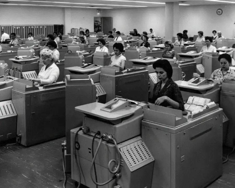 Black and white photo of women working at large, old-fashioned machines in a spacious office. The women are seated in rows, each at their own machine, with papers and cables visible.