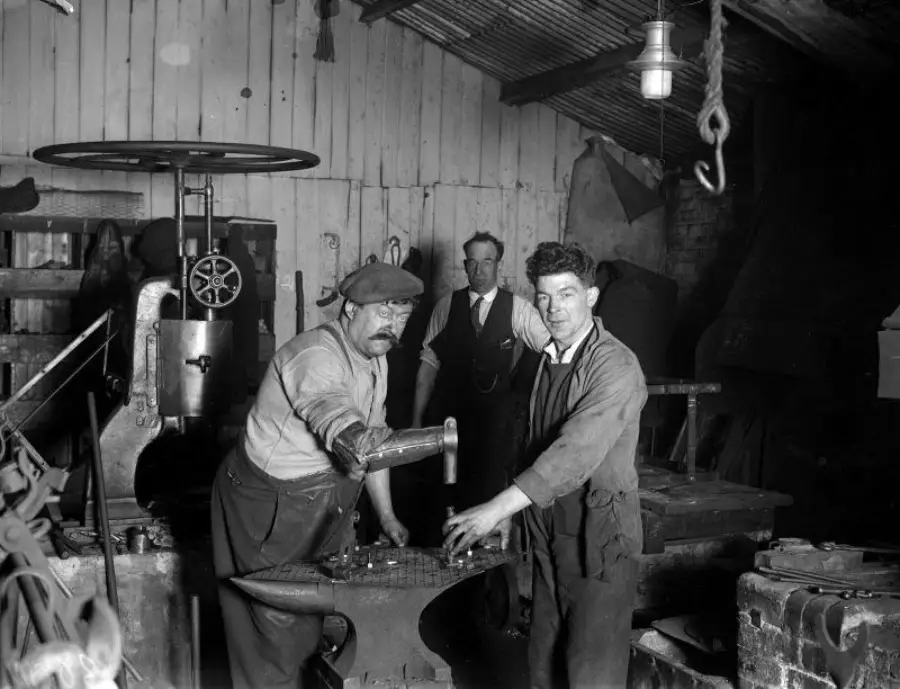 Three men work in a blacksmith workshop beside an anvil and large machinery, surrounded by tools and equipment. The workshop has a corrugated metal roof and wooden walls.