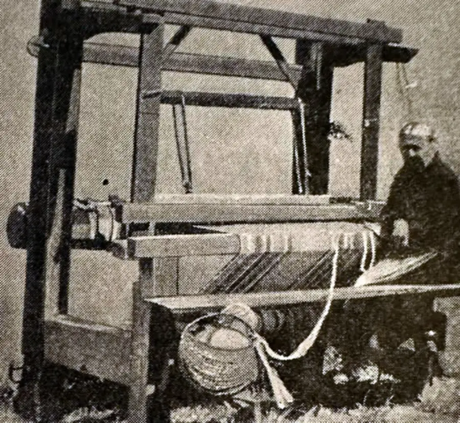 An older person operates a large, traditional wooden loom outdoors. The black-and-white photo shows woven fabric and a basket attached to the loom.