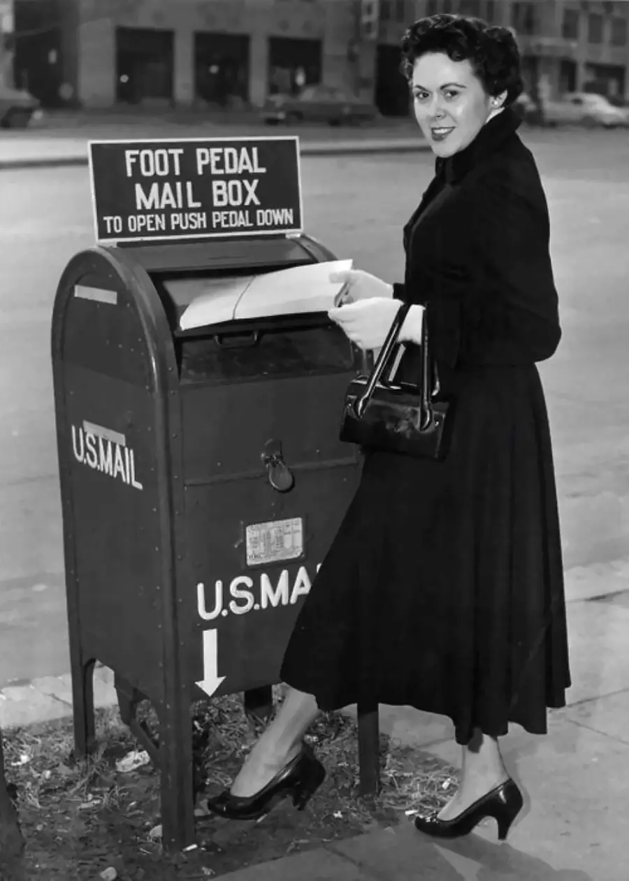A woman in a dark dress uses her foot to open a U.S. Mail mailbox with a pedal while holding letters. The mailbox has a sign explaining the foot pedal operation.