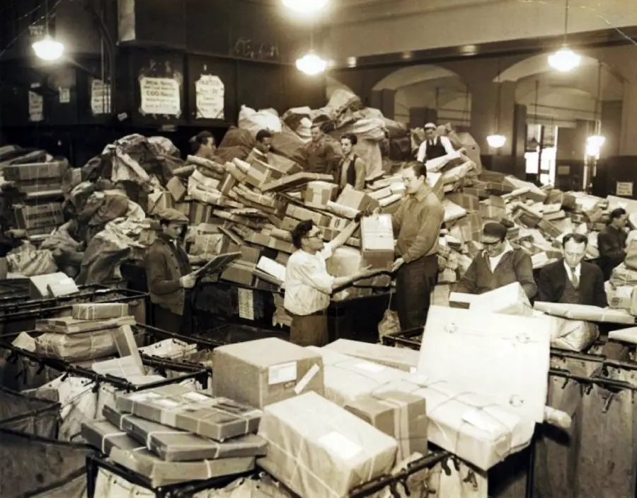 Workers sort and handle a massive pile of packages and mail inside a busy, old-fashioned post office filled with parcels, sacks, and sorting bins.