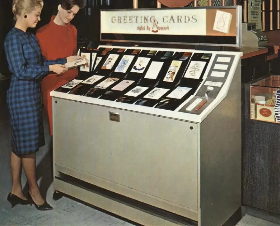 Two women stand at a vintage greeting card vending machine, browsing cards displayed under a sign that reads “Greeting Cards.” The machine has various cards arranged in rows and buttons on the side.