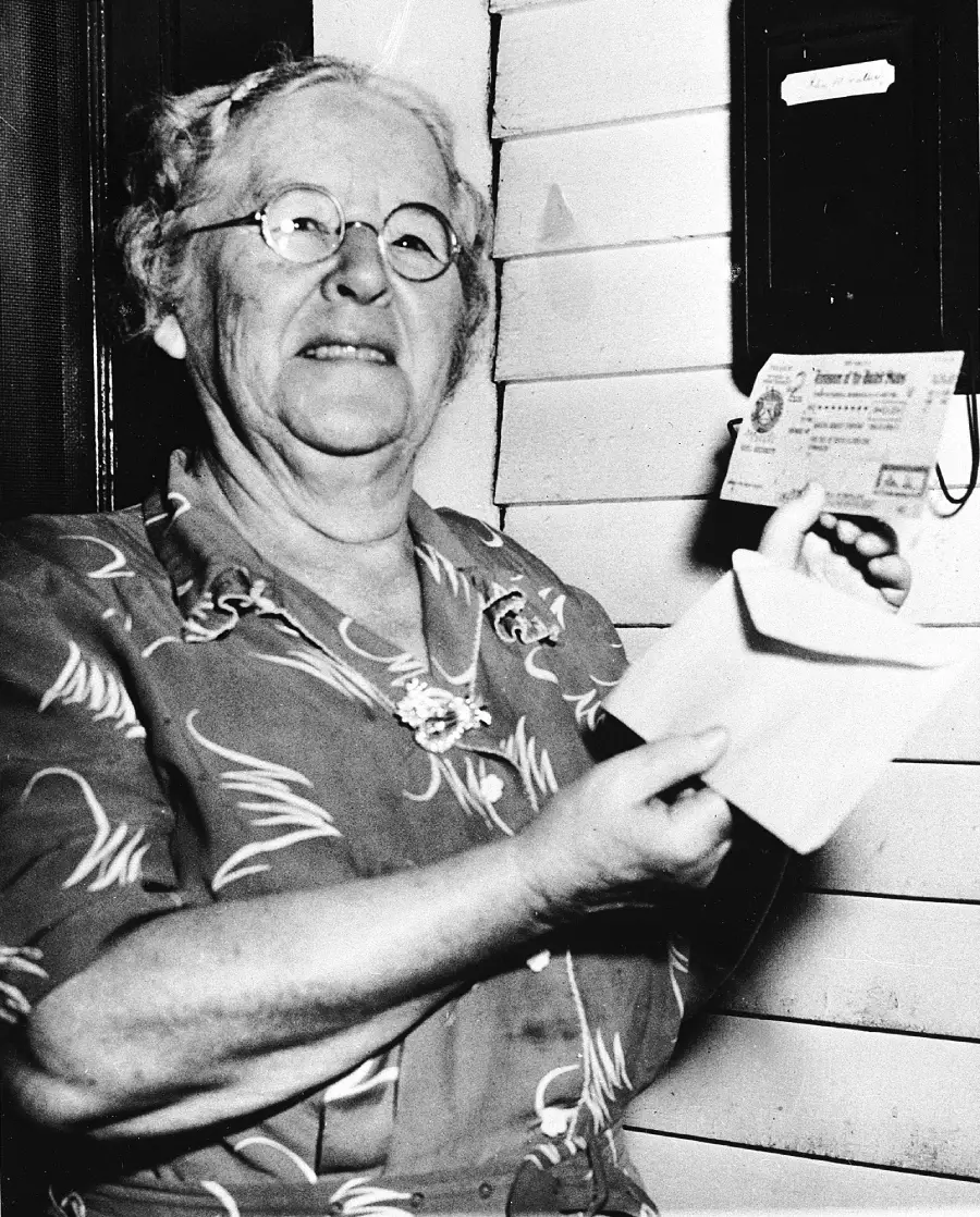 An older woman wearing glasses and a patterned dress smiles while holding an envelope and a letter near a mailbox attached to a wooden wall.
