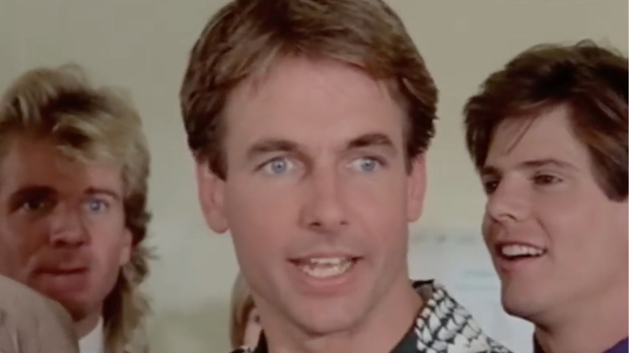 Three men are standing indoors, facing forward. The man in front has short brown hair and blue eyes, speaking animatedly, while the two men behind him look on and smile. The background is out of focus.