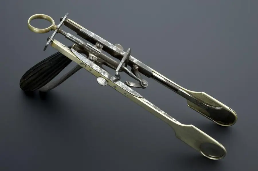 A vintage metal medical instrument with scissor-like handles, a locking mechanism, and two flat, rounded ends, displayed on a dark background.