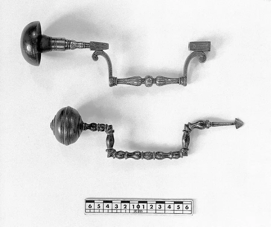 Two ornate, antique metal door handles, each with intricate detailing, are displayed horizontally above a ruler for scale on a plain background. The handles feature decorative knobs and curved elements.
