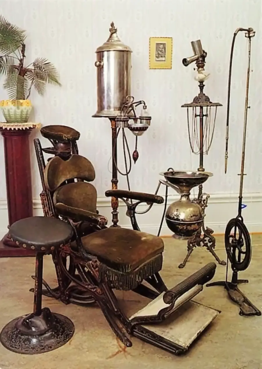 A vintage dentist’s chair with attached footrest, stool, and various old-fashioned dental equipment stands in a room with a potted plant, ornate vase, wallpaper, and a framed picture on the wall.