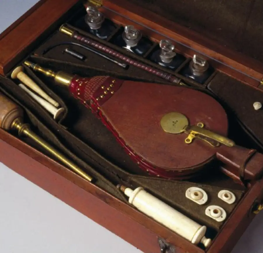 A vintage wooden box containing old medical instruments, including glass vials, a red and brown bellows, rods, and cylindrical tools with brass and white components, neatly arranged in fitted compartments.