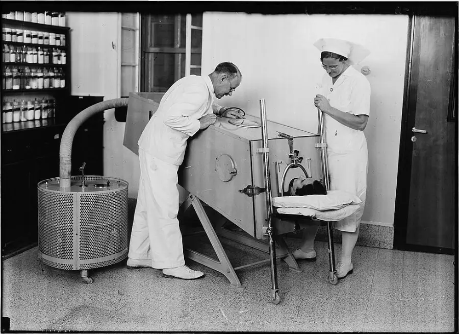 A doctor and nurse examine a patient inside an iron lung machine in a hospital room. Shelves with bottles are visible in the background.