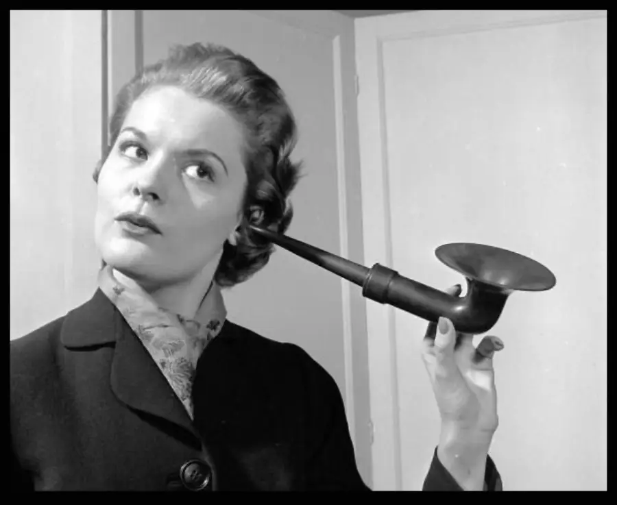 A woman in vintage attire holds an old-fashioned ear trumpet to her ear, looking slightly upward with a curious expression, standing indoors near light-colored doors.