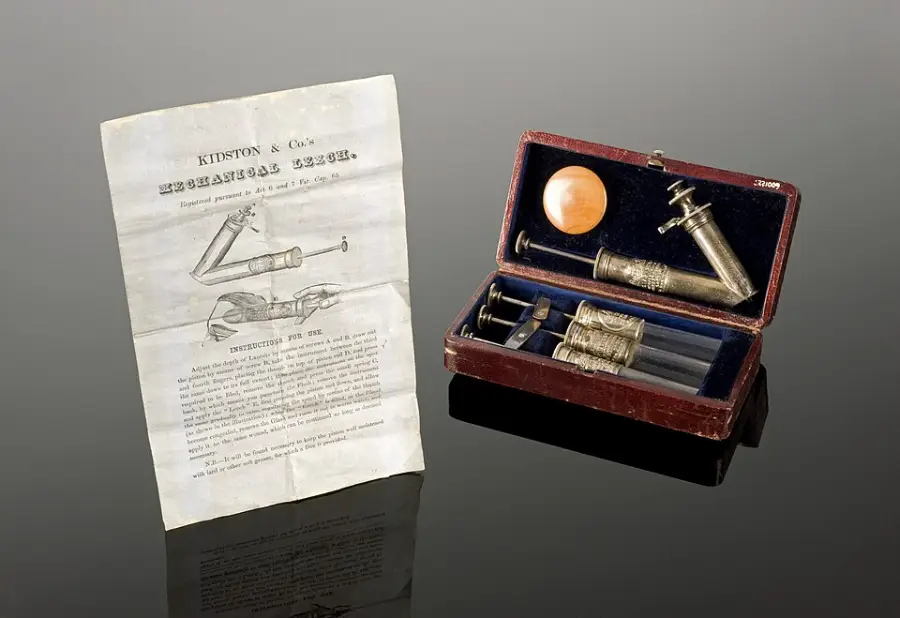 An antique boxed syringe set with two syringes, a needle, and accessories sits next to an illustrated instruction leaflet on a reflective black surface.