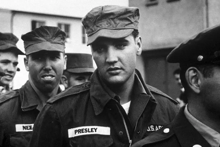 A black and white photo of a man in a military uniform with a name tag reading "Presley," standing among other soldiers. The scene appears to be outdoors, possibly during a military event or gathering.