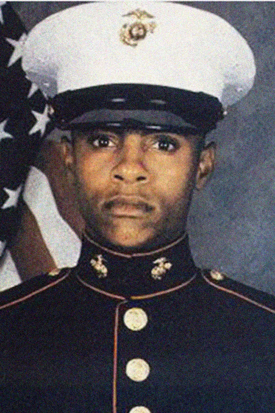 A U.S. Marine in formal dress uniform stands in front of an American flag, looking directly at the camera with a serious expression.
